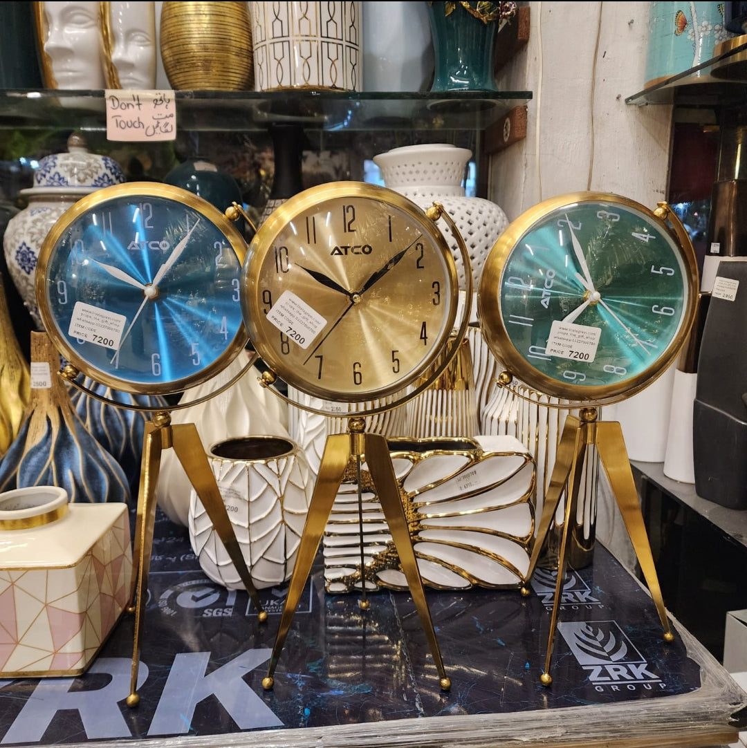 Props ATCO modern metal tripod table clocks featuring a gold-finished frame and stand with blue, gold, and green dials